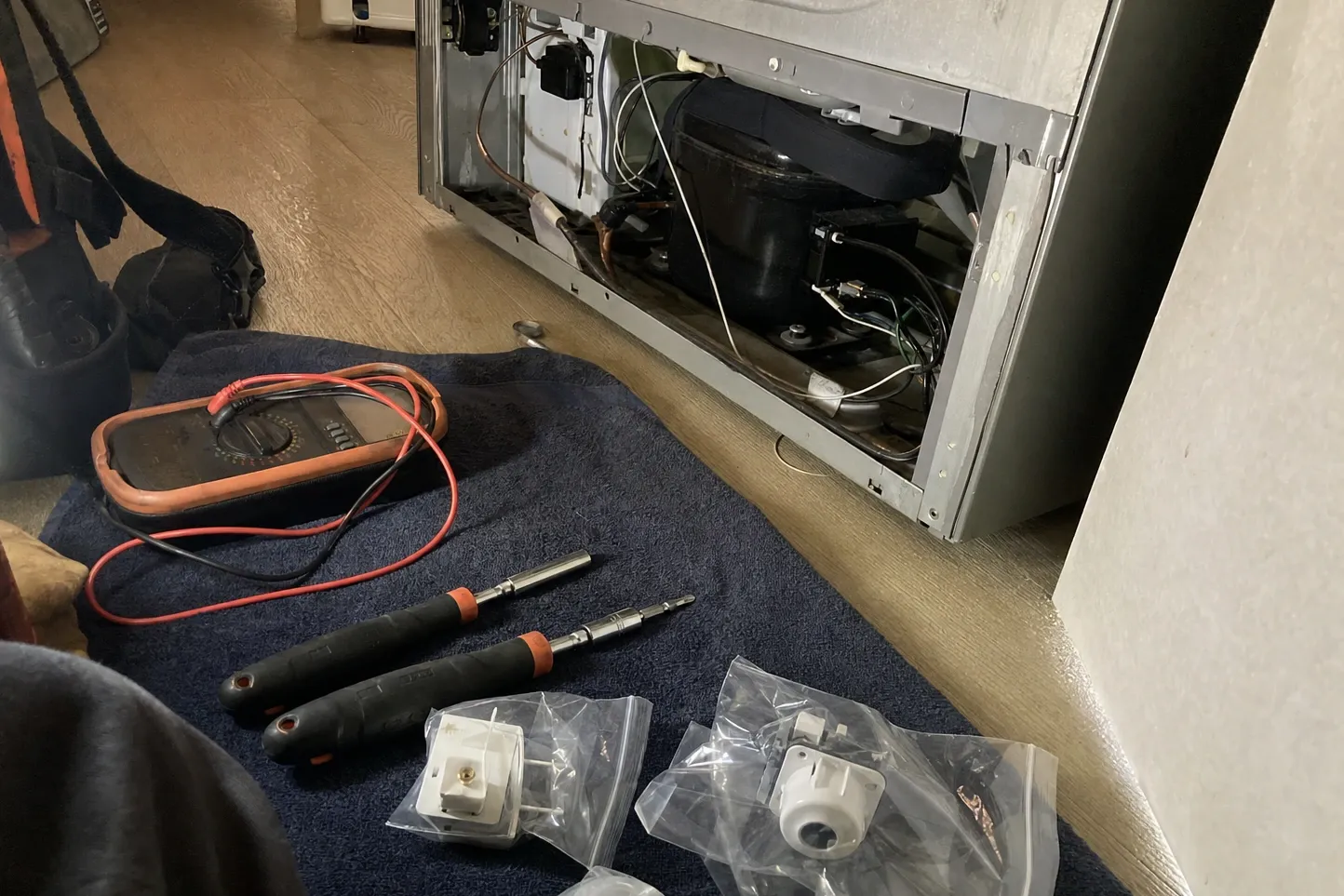 Multimeter, screwdrivers and bagged replacement parts laid out on a towel beside an open machine compartment